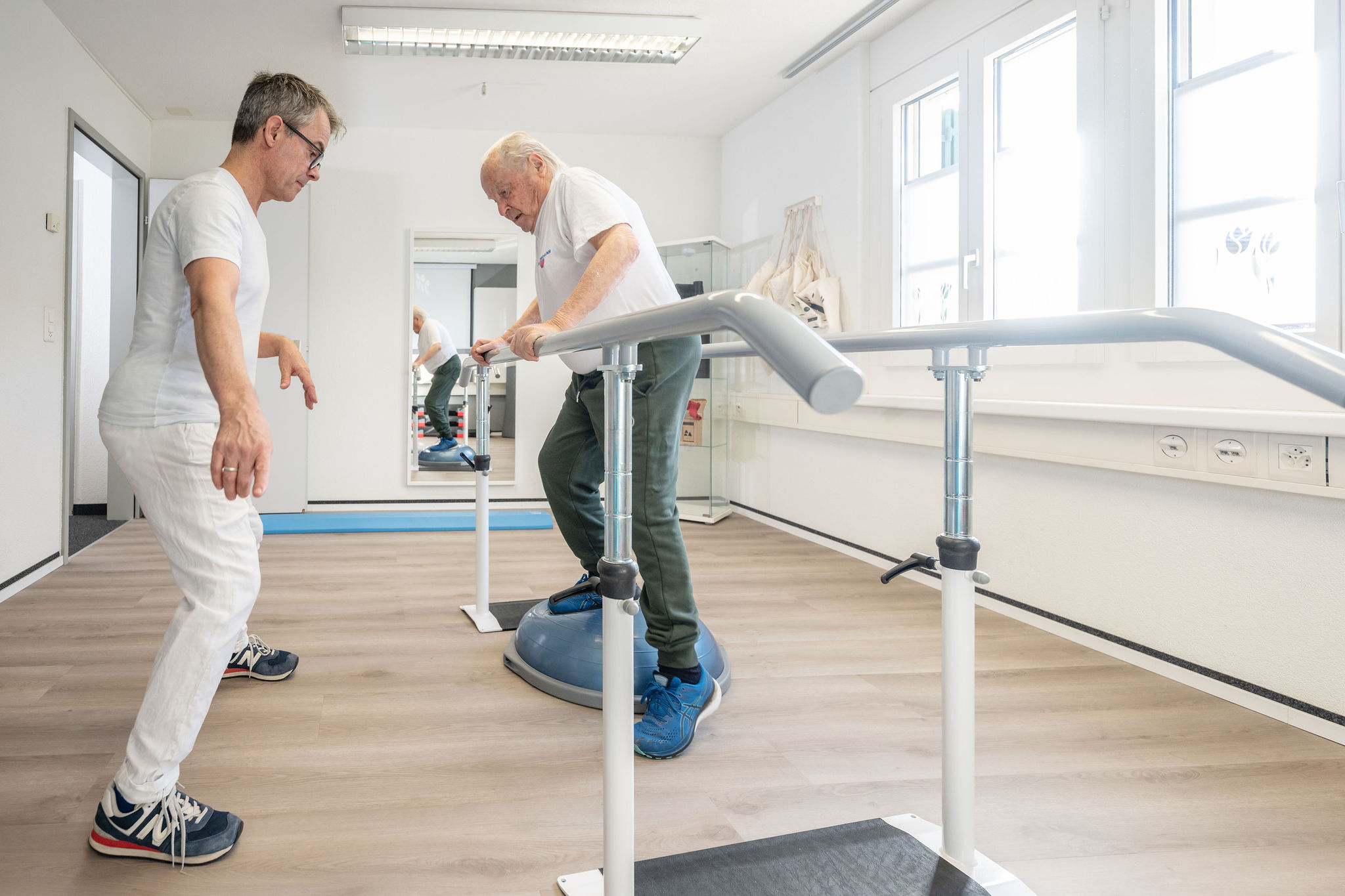 Physiotherapeut Manolito van Haren betreut älteren Patienten beim Gleichgewichtstraining auf dem Bosu-Ball in Physio Kinetic
